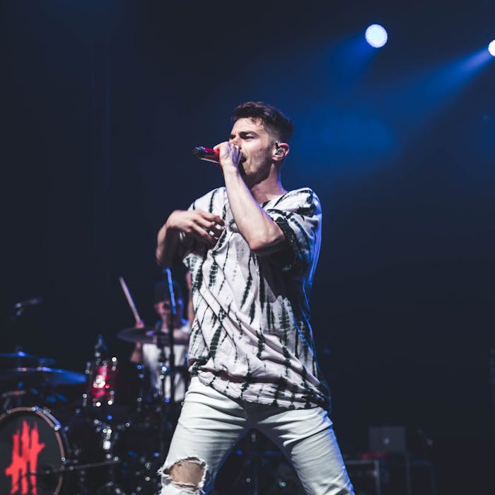 A performer, wearing a tie-dye shirt and ripped jeans, passionately sings into a red microphone on stage. Behind him, a drummer plays in a vibrant, illuminated concert setting featuring Te Pae branding.